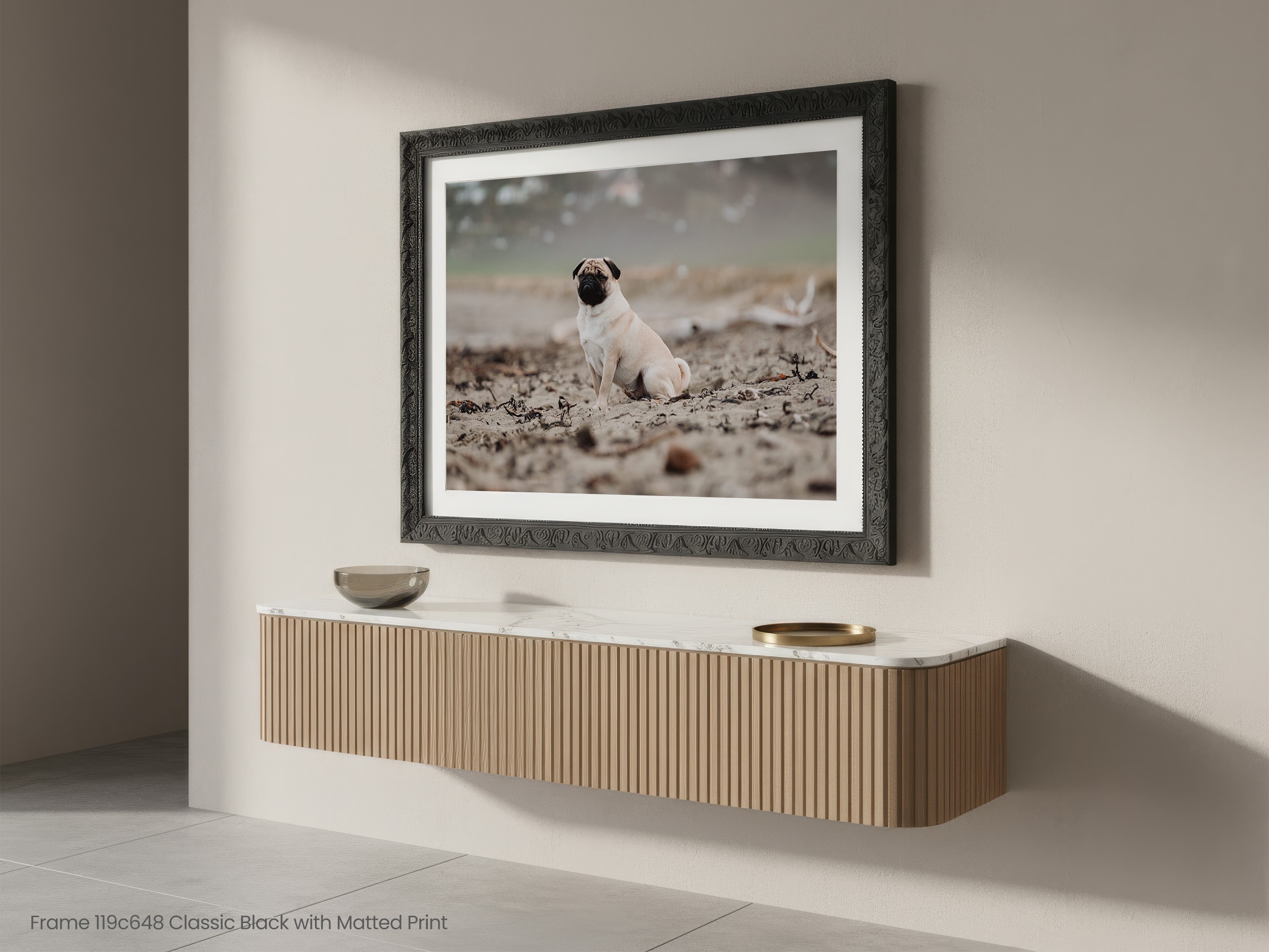 A pug sitting on a beach in a black framed print with a white mat, hanging above a floating sideboard